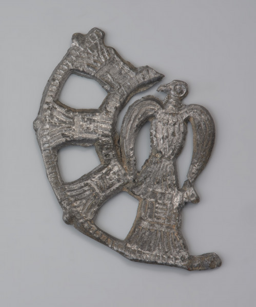 plakietka ażurowa z ptakiem - Ujęcie z przodu. Metalowa, okrągła plakietka ażurowa z wyobrażeniem ptaka w części centralnej. Jego głowa zwrócona jest w lewo. Całość przedstawienia otoczono koncentrycznymi kręgami połączonymi delikatnymi pręcikami. Powierzchnia plakietki jest starannie wyrównana. Na odwrocie znajdują się haczyki do przypinania ozdoby do odzieży.