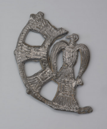 plakietka ażurowa z ptakiem - Ujęcie z przodu. Metalowa, okrągła plakietka ażurowa z wyobrażeniem ptaka w części centralnej. Jego głowa zwrócona jest w lewo. Całość przedstawienia otoczono koncentrycznymi kręgami połączonymi delikatnymi pręcikami. Powierzchnia plakietki jest starannie wyrównana. Na odwrocie znajdują się haczyki do przypinania ozdoby do odzieży.