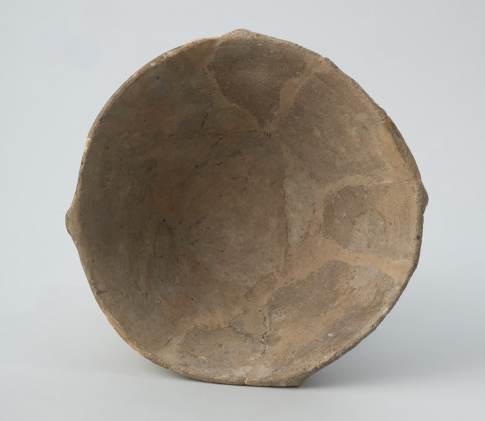 Naczynie gliniane - Ujęcie spodu;  Puchar na pustej nóżce, o klepsydrowatej formie, zdobiony na całej powierzchni ornamentem kreskowo-kłutym wykonanym dwuzębnym narzędziem: na czaszy – w układzie wiszących, wsuwanych kątów, które górą dochodzą do dookolnego pasma linii wykonanych tą samą techniką, a na nóżce – ukośnymi, zbieżnymi pasami linii. Przy krawędzi wylewu znajduje się zdobienie plastyczne w postaci czterech drobnych guzków