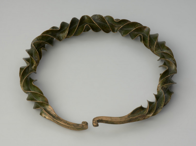 naszyjnik wieńcowaty typu Wendelring - Ujęcie z góry przodu naszyjnika. Naszyjnik formą przypominający wieniec wykonano z brązowego, silnie rozklepanego na krawędziach, czworograniastego pręta o gwiaździstym przekroju, który skręcono (tordowano) wokół własnej osi zmieniając kilkukrotnie kierunek skrętu, uzyskując efekt wąskich, pofałdowanych płaszczyzn o ostrych krawędziach wzdłuż niemal całej długości obręczy. Końce ukształtowane zostały w haczyki służące zapięciu.