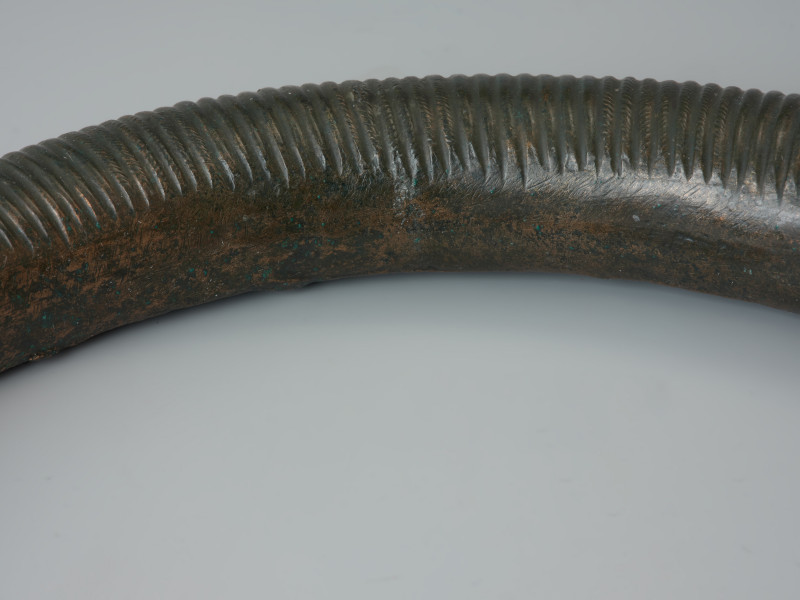 Dwuczęściowy naszyjnik obręczowy - Ujęcie detalu z przodu. Zapinany haczykami naszyjnik z brązu, złożony z dwóch obręczy, zdobiony poprzecznym żebrowaniem oraz motywem zygzaka.