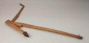 radło płozowe krzywogrządzielowe - Ujęcie przodu radła z góry. Radło z prostokatnym otworem na trzonek i rączką równoległą do płaszczyzny płozy. Przedmiot wykonano z naturalnie rozgałęzionego fragmentu drzewa, prawdopodobnie brzozowego. Część górną rozwidlenia tworzy grządziel (dyszel) służąca do zaprzężenia bydła pociągowego. Dolny człon rozgałęzienia stanowi płoza z piętką będąca elementem pracującym po powierzchni ziemi. Grządziel oraz płozę wzmacnia słupica łącząca obydwa elementy. Ponadto radło ma prostokątny otwór z osadzonym dębowym trzonkiem, na końcu którego uformowano równoległą do płaszczyzny płozy rączkę.