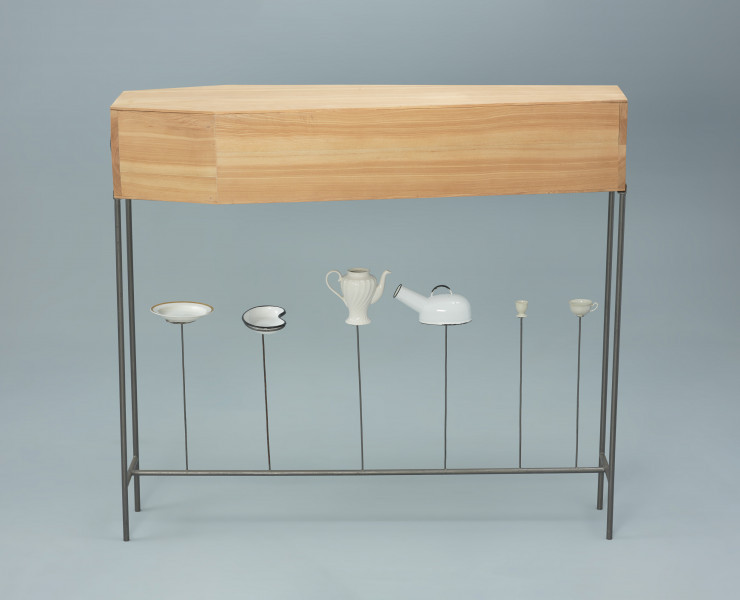 obiekt Utensylia - Ujęcie z przodu w innym układzie. Mobilny obiekt z pudłem w kształcie trumny wykonanym z jasnego drewna. Pudło posadowione na zakończonym kółkami metalowym, czarnym stelażu. W bocznych, szczytowych ścianach pudła okrągłe soczewki optyczne. Na stelażu, pod pudłem sześć pionowych prętów z elementami wyposażenia sanitarnego i zastawy stołowej. Kolejno od czoła obiektu: talerz, nerka medyczna, dzbanek, kaczka sanitarna, kieliszek na jajko i filiżanka.