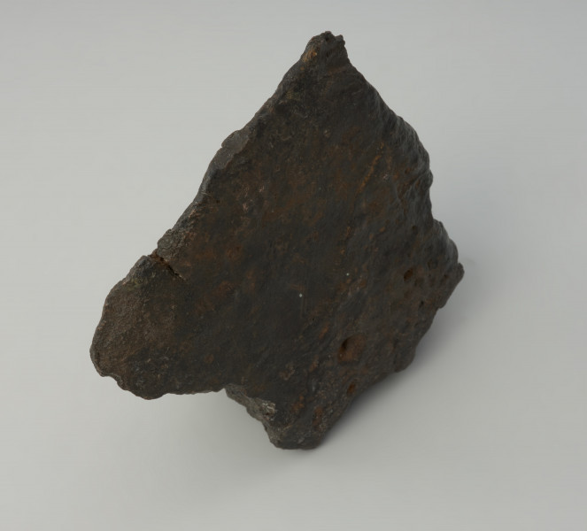 bryła metalu - Ujęcie w pionie z góry. Fragment placka odlewniczego o nieregularnym, czworobocznym kształcie z zachowanym jednym z pierwotnych boków o ostrej krawędzi. W przekroju poprzecznym trójkątny.