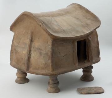 naczynie sepulklarne, urna domkowa - Ujęcie z przodu skosem w prawo z otwartym wejściem. Urna gliniana w formie domku na czterech profilowanych filarach, z wejściem w jednej z dłuższych ścian, zdobiona poziomymi i pionowymi liniami rytymi.