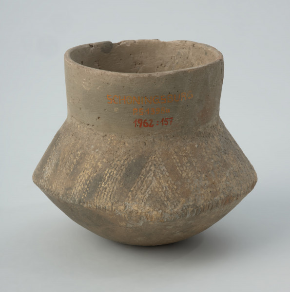 Naczynie gliniane - Ujęcie z tyłu; Naczynie wazowate o dwustożkowatym brzuścu, pionowej szyjce i niewyodrębnionym dnie. Brzusiec o zarysie czworobocznym w rzucie z góry. Zdobione w górnej części brzuśca ukośnymi pasmami złożonymi z 3 podwójnych linii kreskowo-kłutych wykonanych dwuzębnym narzędziem. Pasma trzykrotnie zmieniają kierunek nachylenia, tworząc układ krokwiasty. Cały układ podkreślony dookolnym pasmem odcisków paznokcia na załomie brzuśca. Szyjka naczynia, niemal w całości zrekonstruowana, prawdopodobnie nie była zdobiona, podobnie jak dół brzuśca