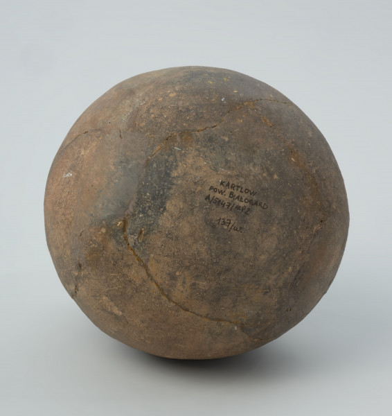 Naczynie gliniane - Ujęcie spodu.  Amfora o baniastym brzuścu i cylindrycznej szyjce z parą taśmowatych uch umieszczonych pomiędzy nasadą szyjki i górną partią brzuśca, zdobiona na szyjce pod wylewem podwójnym pasmem pionowych stempelków (słupków), analogicznym, ale pojedynczym pasmem u nasady szyjki, a w górnej części brzuśca dookolnie parami pionowych linii odcisków sznura. Naczynie zrekonstruowane i uzupełnione gipsem barwionym. Barwa od szarobrunatnej przez brunatną po brązową.