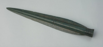 Grot oszczepu lub włóczni typu Ullerslev - Ujęcie w poziomie przodu grotu. Długi, smukły grot o któtkiej tulejce, wzmocniony żeberkiem. Tulejka zdobiona jest przy wylocie trzema pasmami zygzakowymi, poprzedzielanymi pasmami dookolnych linii, całość wieńczą trójkąty. Zabytek pokrywa zielona patyna.