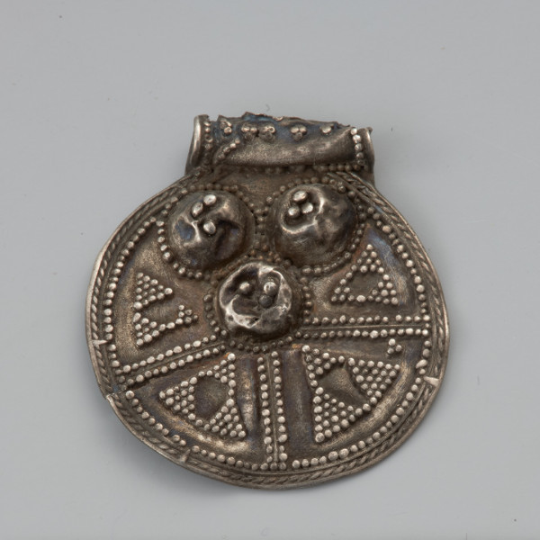 ozdoba; biżuteria; zawieszka - Ujęcie przodu zawieszki uszkiem do góry. Okrągła zawieszka wycięta ze srebrnej blachy o średnicy 2,5 cm, wraz z pasmem zawiniętym w rurkowate uszko. Zdobiona jest po jednej stronie trzema guzkami, z których każdy jest otoczony wieńcem przylutowanych półkolistych granulek. Również na każdym z guzków znajdują się po trzy granulki. Na pozostałej powierzchni blaszki znajdują się cztery promieniście rozłożone pola wypełnione ornamentem trójkątów wykonanym w technice granulacji oraz łańcuszkiem półjodełki. Uszko, również zdobione jest granulkami. Do tylnej, niezdobionej strony, od boków uszka do około połowy wysokości okrągłej części, przylutowane są wąskie pasemka blachy wzmacniające zawieszkę. Zarówno uszko jak i zawieszka właściwa poniżej linii guzków są pęknięte.