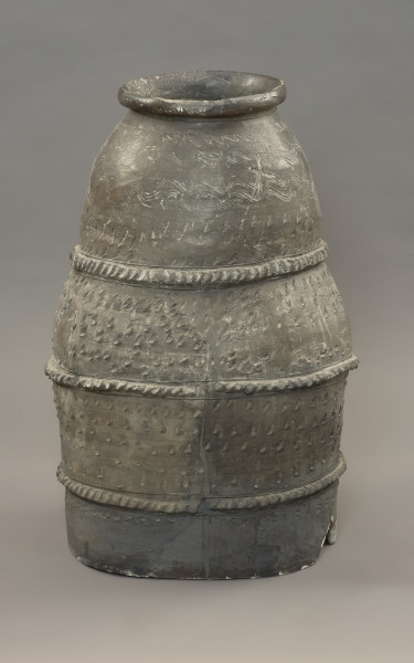 pojemnik; naczynie zasobowe - Ujęcie z lewej strony. Częściowo zrekonstruowane naczynie zasobowe o workowatym kształcie. Powierzchnia naczynia jest ornamentowana czterema strefami z motywami zdobniczymi oddzielonymi trzema dookolnymi listwami plastycznymi, na których znajdują się szerokie ukośne nacięcia przypominające karby. Pierwszą strefę przyozdabiają cztery rzędy niestaranie wykonanych, dookolnych linii falistych. Tuż pod falami znajdują się trzy rzędy lewo-skośnych nacięć. Drugą i trzecią strefę zdobią po cztery rzędy lewo-skośnych nacięć. Zasobowiec wylepiono z glinianych wałków z masy garncarskiej wzbogaconej domieszką piasku oraz granitu o różnorodnym stopniu granulacji. Naczynie wylepiono najprawdopodobniej ręcznie bez użycia koła garncarskiego.