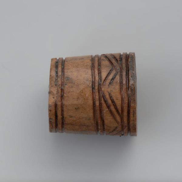 oprawka stożkowata - Ujęcie prawej strony oprawki w poziomie. Oprawka ma stożkowaty kształt o ściętym wierzchołku. Ślady pozostawione na jej powierzchni świadczą o wykorzystaniu do jej produkcji co najmniej trzech narzędzi – piły, noża oraz świdra. Na powierzchni oprawki znajduje się mało skomplikowany wzór zdobniczy o motywie liniowym naniesionym za pomocą ostrego narzędzia. Górną część zajmuje ornament w postaci dwóch dookolnych linii rytych. Natomiast na przeciwległej stronie występuje ozdobnik w formie linii zygzakowatych wpisanych od góry i od dołu w podwójne linie ryte.