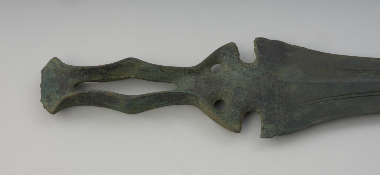 miecz ze sztabą do rękojeści - Ujęcie głowni miecza w poziomie z tyłu. Zachowany w całości miecz obosieczny o smukłej głowni z widocznymi podwójnymi struzinami na powierzchni. Sztaba do rękojeści ma oryginalną formę, poszerzoną pośrodku i zwieńczoną wachlarzowatym zakończeniem. Dodatkowo na całej jej długości przebiega charakterystyczna, rombowata szczelina. Pókolista podstawa sztaby zaopatrzona jest w 2 otwory na nity. Obydwie powierzchnie miecza są lekko porowate i pokryte zieloną patyną.