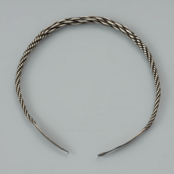 naszyjnik srebrny - Ujęcie tyłu naszyjnika z góry. Srebrny naszyjnik spleciony z pięciu par drutów owiniętych wokół siebie, z końcami rozklepanymi w owalne, niezdobione płytki. Jedna z nich zakończona została haczykiem, końcówka drugiej jest ułamana. W części środkowej druty są skręcone luźniej, co powoduje, ze obręcz jest w tym miejscu grubsza, natomiast po obu stronach znajdują się fragmenty, gdzie splot jest nierówny.