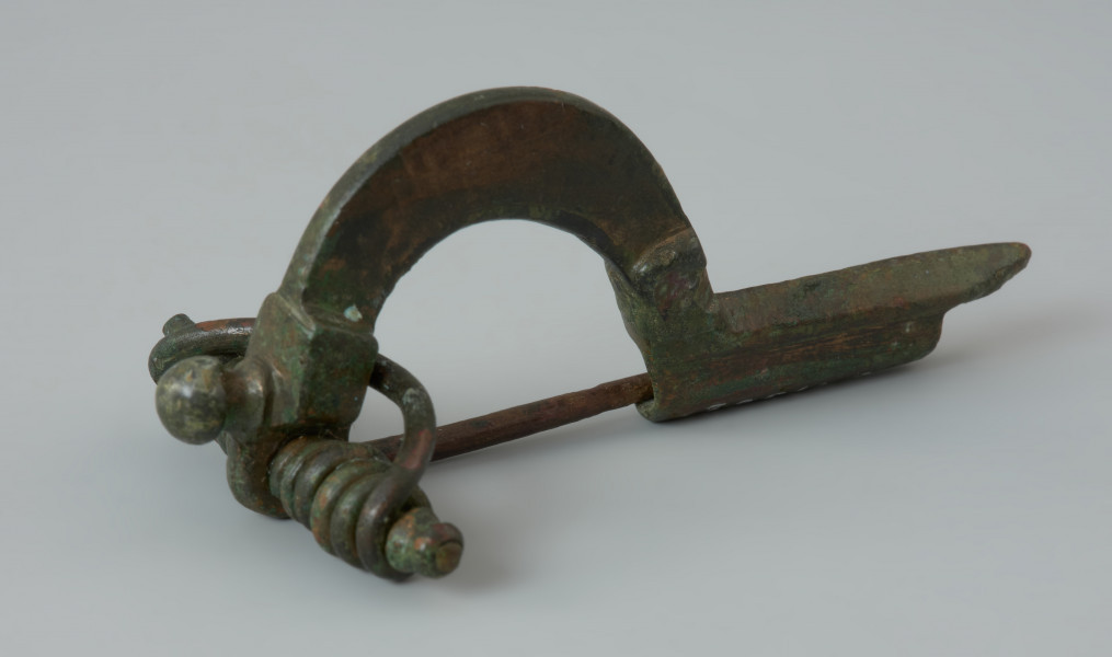 Fibula z brązu z profilowanym guzkiem na główce - Ujęcie z przodu. Fibula z brązu, zwana również zapinką. Razem z grzebieniem rogowym stanowiła element wyposażenia grobu szkieletowego.