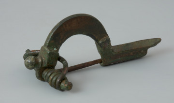 Fibula z brązu z profilowanym guzkiem na główce - Ujęcie z przodu. Fibula z brązu, zwana również zapinką. Razem z grzebieniem rogowym stanowiła element wyposażenia grobu szkieletowego.