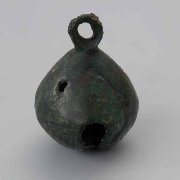 zawieszka dzwoneczkowata z Wolina - Ujęcie tyłu zawieszki w pionie. Zawieszka dzwoneczkowata wykonana z brązowej blachy, spatynowanej na ziolono. Korpus przypomina kształtem pustą w środku kroplę rozciętą u dołu na krzyż z jednym ramieniem dłuższym i szerszym od drugiego. Dodatkowo dolna powierzchnia jest zdobiona żłobkowaniem. W rzucie z góry zawieszka ma kształt zbliżony do rąbu, u góry posiada otwór z trójkątnego w przekroju drutu. W jej wnętrzu znajduje się brązowa kulka. Powierzchnia dzwoneczka jest lekko zniszczona, a w ściance znajduje się niewielki ubytek.