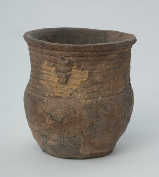 Naczynie gliniane - Ujęcie z tyłu; Puchar o kształcie esowatym, być może pierwotnie zaopatrzony w ucho, zdobiony na szyjce dookolnym pasmem złożonym z 10 rzędów ornamentu odcisków sznura. Naczynie zrekonstruowane na podstawie drobnych fragmentów wylewu, szyjki, brzuśca i dna, uzupełnione barwionym gipsem. Powierzchnia szarobrunatna, z powierzchnią niedokładnie wyrównaną. W masie, na powierzchni widoczna domieszka mineralna.