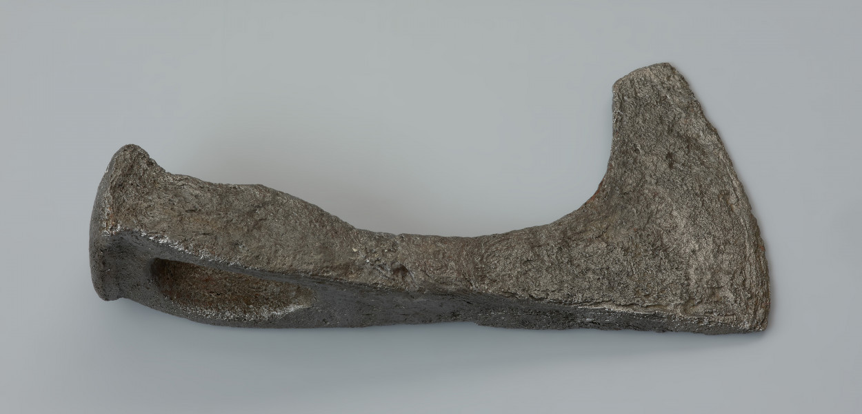 topór bojowy (czekan) - Ujęcie z góry lewej strony z góry. Żeleźce jest masywne, z ostrzem lekko asymetrycznie opuszczonym w stosunku do toporzyska. Obuch jest zakończony krótkim, silnie wyodrębnionym, sztabkowatym zgrubieniem, tak zwanym młotkiem. Szyjka przechodzi w wąskie, asymetryczne i łukowate ostrze zakończone wyodrębnioną, ukośnie podciętą wąską brodą.