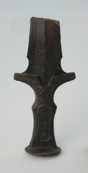broń, miecz - Ujęcie całej rękojeści i głowicy z przodu w pionie. Brązowy miecz z czworokatną, dekorowaną żłobieniami rękojeścią, głowicą w formie owalnej tarczki oraz jelcem ze skrzydełkami i półkolistym wycięciem.