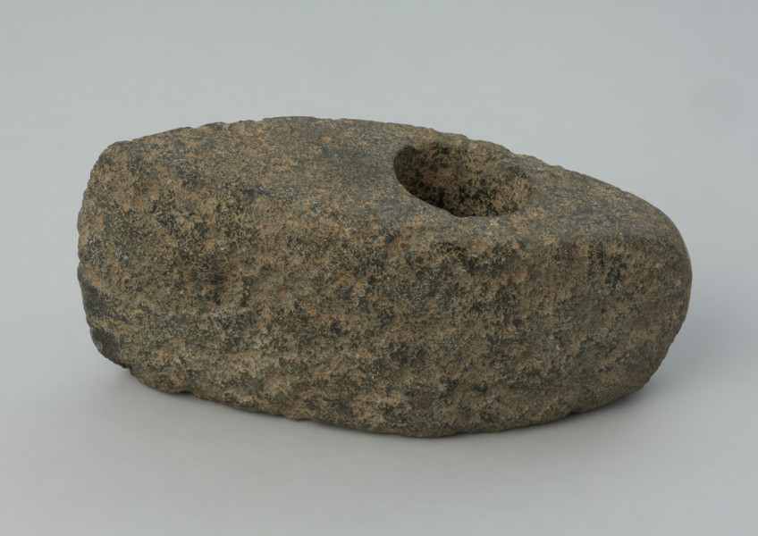 Topór kamienny - Ujęcie z przodu; Topór o wydłużonej części tylnej zakończonej wąskim obuchem, z wywierconych otworem przesuniętym ku tyłowi. Przekrój poprzeczny czworokątny wysoki. W w rzucie bocznym trapezowaty z lekko skośnym ostrzem i lekko łukowatym obuchem. Przy otworze, na stronie górnej, ślady wykucia płytkiej miseczki pod wiercenie. Powierzchnia zerodowana ze śladami szlifowania i puncowania. Ostrze wtórnie uszkodzone.