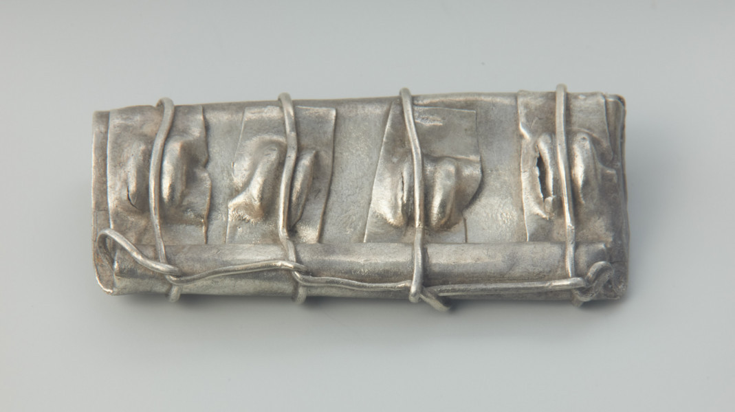 Kaptorga - Ujęcie przodu kaptorgi z góry.  Kaptorga, prostokątna zawieszka ukształtowana ze srebrnej blachy zagiętej wpół, zawiniętej z jednej strony w długie, rurkowate uszko, pod którym jest przylutowany drugi koniec blachy. Jeden bok jest zamknięty poprzez zagięcie na siebie bocznych krawędzi blachy, drugi natomiast pozostawiono otwarty. Do przedniej ścianki kaptorgi przymocowane zostały cztery prostokątne, pionowo ułożone, blaszki z wytłoczonymi guzkami. Całość oplata drucik przechodzący przez każdy guzek i biegnący wzdłuż rurkowatego uszka. Przedmiot został odkryty w 1933 roku w Dobrociechach w nieznanych okolicznościach i pochodzi ze skarbu monet i ozdób srebrnych