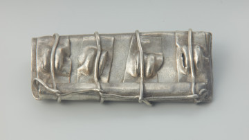 Kaptorga - Ujęcie przodu kaptorgi z góry.  Kaptorga, prostokątna zawieszka ukształtowana ze srebrnej blachy zagiętej wpół, zawiniętej z jednej strony w długie, rurkowate uszko, pod którym jest przylutowany drugi koniec blachy. Jeden bok jest zamknięty poprzez zagięcie na siebie bocznych krawędzi blachy, drugi natomiast pozostawiono otwarty. Do przedniej ścianki kaptorgi przymocowane zostały cztery prostokątne, pionowo ułożone, blaszki z wytłoczonymi guzkami. Całość oplata drucik przechodzący przez każdy guzek i biegnący wzdłuż rurkowatego uszka. Przedmiot został odkryty w 1933 roku w Dobrociechach w nieznanych okolicznościach i pochodzi ze skarbu monet i ozdób srebrnych