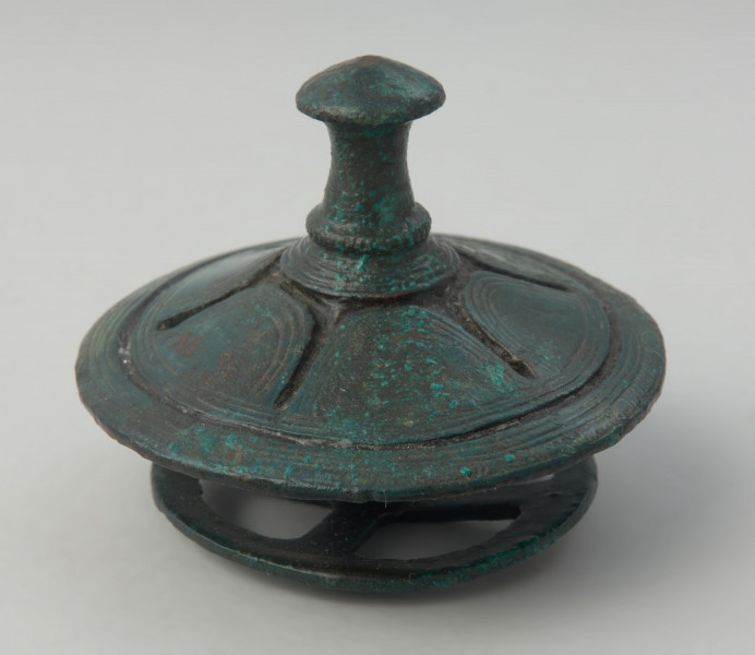 Brązowy tutullus złożony z dwóch tarczek oraz kolca. - Ujęcie totullusa z boku. Brązowy tutullus złożony z dwóch tarczek oraz kolca. Górna tarczka posiada  zdobienie w postaci żłobków, ażuru, linii rytych, nacięć oraz motywu solarnego. Dolna tarczka ma formę koła z czterema szprychami.