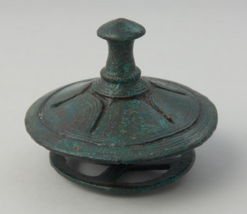 Brązowy tutullus złożony z dwóch tarczek oraz kolca. - Ujęcie totullusa z boku. Brązowy tutullus złożony z dwóch tarczek oraz kolca. Górna tarczka posiada  zdobienie w postaci żłobków, ażuru, linii rytych, nacięć oraz motywu solarnego. Dolna tarczka ma formę koła z czterema szprychami.