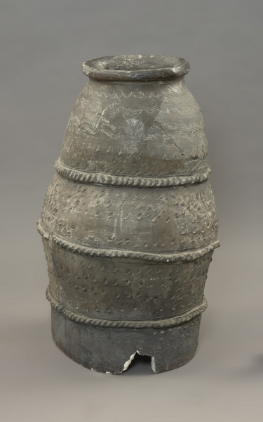 pojemnik; naczynie zasobowe - Ujęcie z tyłu. Częściowo zrekonstruowane naczynie zasobowe o workowatym kształcie. Powierzchnia naczynia jest ornamentowana czterema strefami z motywami zdobniczymi oddzielonymi trzema dookolnymi listwami plastycznymi, na których znajdują się szerokie ukośne nacięcia przypominające karby. Pierwszą strefę przyozdabiają cztery rzędy niestaranie wykonanych, dookolnych linii falistych. Tuż pod falami znajdują się trzy rzędy lewo-skośnych nacięć. Drugą i trzecią strefę zdobią po cztery rzędy lewo-skośnych nacięć. Zasobowiec wylepiono z glinianych wałków z masy garncarskiej wzbogaconej domieszką piasku oraz granitu o różnorodnym stopniu granulacji. Naczynie wylepiono najprawdopodobniej ręcznie bez użycia koła garncarskiego.