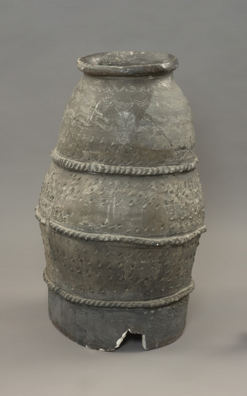 pojemnik; naczynie zasobowe - Ujęcie z tyłu. Częściowo zrekonstruowane naczynie zasobowe o workowatym kształcie. Powierzchnia naczynia jest ornamentowana czterema strefami z motywami zdobniczymi oddzielonymi trzema dookolnymi listwami plastycznymi, na których znajdują się szerokie ukośne nacięcia przypominające karby. Pierwszą strefę przyozdabiają cztery rzędy niestaranie wykonanych, dookolnych linii falistych. Tuż pod falami znajdują się trzy rzędy lewo-skośnych nacięć. Drugą i trzecią strefę zdobią po cztery rzędy lewo-skośnych nacięć. Zasobowiec wylepiono z glinianych wałków z masy garncarskiej wzbogaconej domieszką piasku oraz granitu o różnorodnym stopniu granulacji. Naczynie wylepiono najprawdopodobniej ręcznie bez użycia koła garncarskiego.