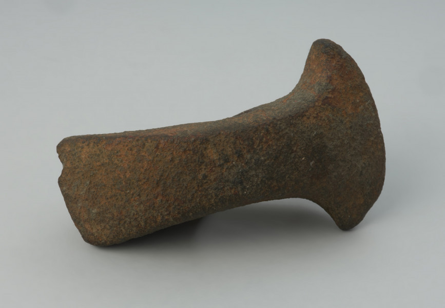 Topór kamienny - Ujęcie z przodu; Topór z szerokim, symetrycznie rozszerzonym ostrzem i nieznacznie rozszerzonym obuchem, o prostokątnym przekroju poprzecznym, wykonany z szarego surowca skalnego. Otwór niemal kolisty, cylindryczny. Powierzchnia silnie zerodowana.