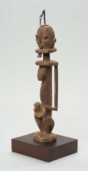 rzeźba - Ujęcie z przodu z lewej strony. Drewniana, rzeźbiona Postać z dzieckiem na kolanach. Siedzi w pozycji przykucniętej. Głowa i tułów nieproporcjonalnie duże w stosunku do nóg. Oczy, nos, usta, uszy mocno wypukłe. Dziecko ułożone na kolanach, zasłania twarz rękoma. Na głowie fragment żelaza.