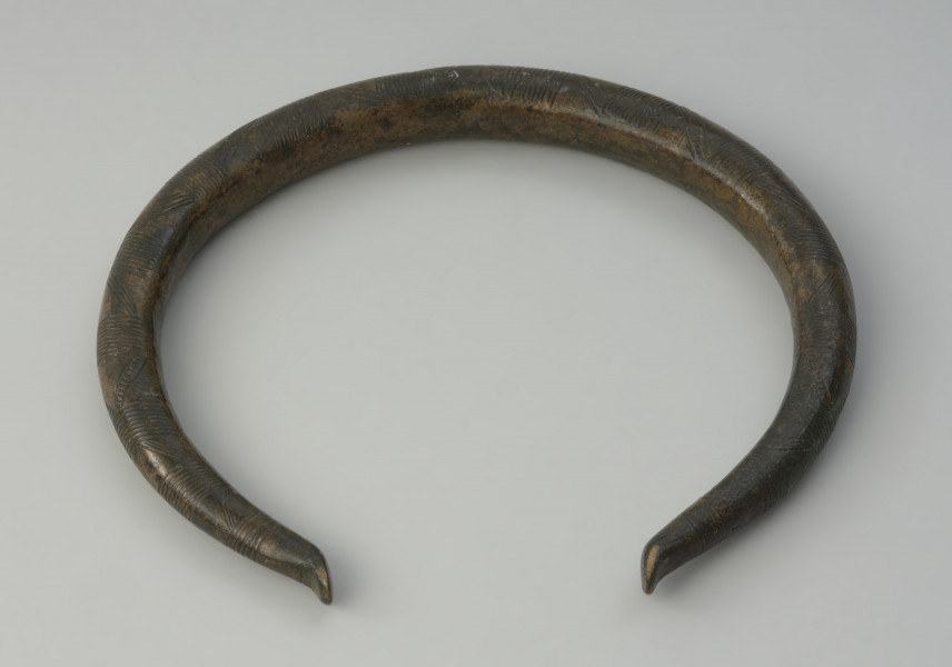 Brązowy naszyjnik - Ujęcie z góry tyłu naszyjnika. Brązowy naszyjnik wykonany z grubego okrągłego pręta. Jego zakończenia są odgięte na zewnątrz. Zdobiony jest poprzecznymi kreskami, trójkątami oraz łukami.