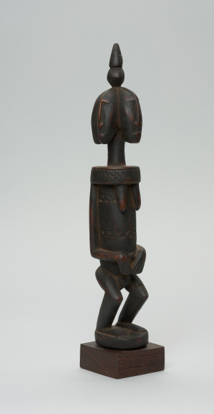 rzeźba - Ujęcie z przodu z prawej strony. Drewniana, rzeźbiona postać z wyraźnie zaznaczonymi piersiami, a także z czterema głowami. Każda głowa zwrócona jest w inna stronę. Na czubku głowy stożkowata bryła. Twarze zaznaczone bardzo symbolicznie, nos w kształcie strzałki. Ramiona od szyi oddzielone prostą linią. Ręce przylegające do tułowia, dłonie złożone na łonie/podbrzuszu. Nogi krótkie, podkurczone w kolanach. Na brzuchu, na wysokości pępka dekoracyjne trójkątne nacięcia.