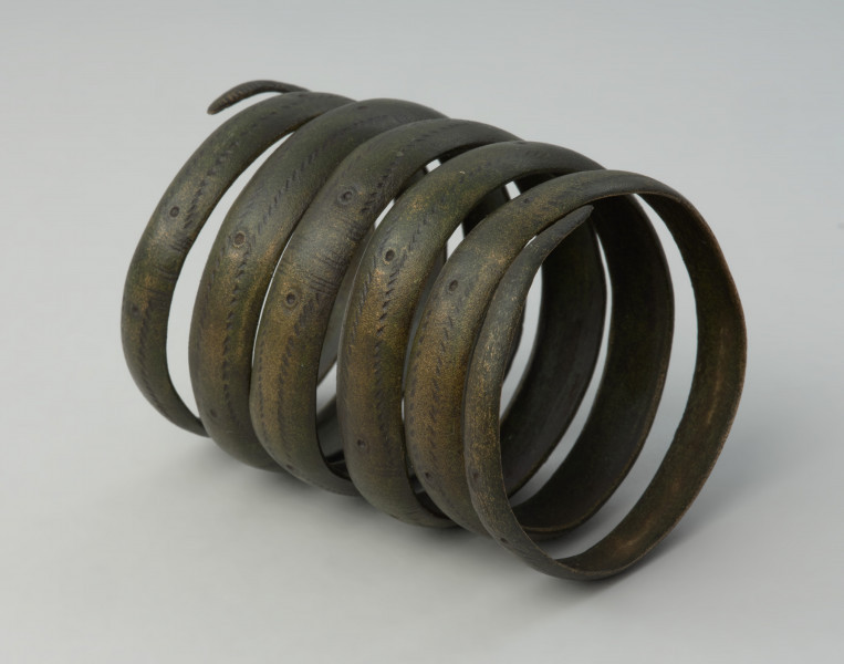 naramiennik spiralny typu kujawskiego - Ujęcie w poziomie skosem w prawo. Sześciozwojowy naramiennik wykonany z wąskiej brązowej taśmy. Powierzchnię taśmy zdobi ornament oczkowy połączony z ukośnymi nacięciami i poprzecznymi żłobkami. Starannie opracowane zakończenia naramiennika są uformowane w szpic.