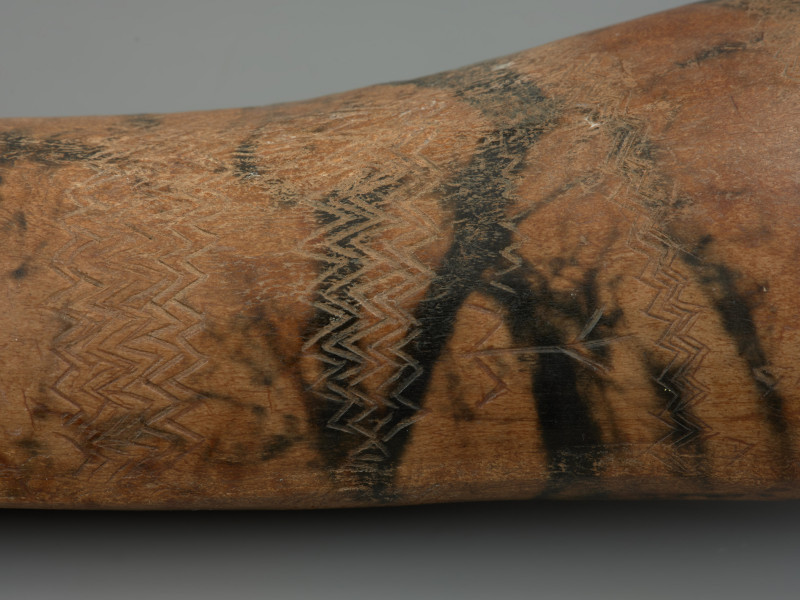 insygnium z Rusinowa - ujęcie detalu z drugiej strony; Bogato ornamentowane insygnium z poroża łosia. Zabytek pokryty jest ornamentem, na który składają się serie poprzecznych zygzaków, zgrupowanych w siedem pasm z jednej strony przedmiotu i osiem z drugiej. Pasma te zawierają od jednej do jedenastu linii. Powierzchnia jest wyświecona na skutek wielokrotnego owijania w skórę. W liniach rytych znajduje się ciemna substancja, prawdopodobnie brud, który nagromadził się w trakcie funkcjonowania zabytku. Na tej stronie znajdują się ciemne, liniowe odbarwienia będące efektem kontaktu z korzeniami roślin.