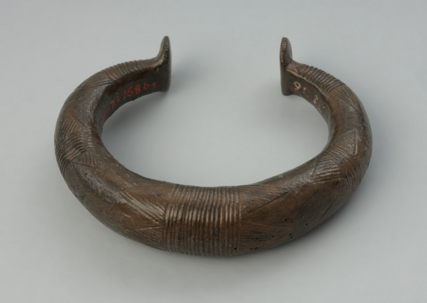 Bransoleta obręczowa - Ujęcie z góry przodu bransolety. Masywna bransoleta obręczowa o owalnym kształcie, odlana z brązu w formie grubego pręta. Jej zakończenia mają kształt niewielkich stopek. Powierzchnię obręczy zdobią grupy poprzecznych kresek oraz motyw tzw. wilczych zębów.