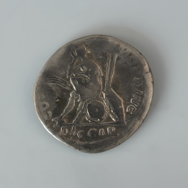 Ujęcie rewersu. Denar Trajana. Powierzchnie są w znacznym stopniu starte, a wyobrażenia i napisy są częściowo nieczytelne. Na awersie widnieje popiersie cesarza w wieńcu laurowym, zwrócone prawym profilem, na lewym dalszym barku widoczna udrapowana szata. W otoku znajduje się napis IMP TRAIANO AVG GER DAC PM TRP. Na rewersie znajduje się personifikacja Dacii w żałobie lub wyobrażenie pokonanego Daka. Jest zwrócona w prawo postać w stroju dackim: spodniach, krótkiej tunice (płaszczu?) i szpiczastej czapce. Postać ta siedzi na stosie uzbrojenia prawa dłonią zakrywa twarz. Wokół wizerunku znajduje się częściowo tylko zachowany napis COS V PP SPQR OPTIMO PRINC.