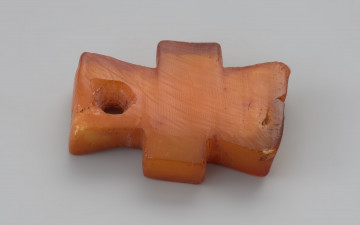 zawieszka krzyżykowata - Ujęcie prozdu zawieszki z góry w poziomie. Zawieszka bursztynowa w kształcie krzyżyka nieznacznie nieforemnego w kolorze mlecznopomarańczowym, miejscami przejrzystym. Ramiona pionowe rozszerzają się na zewnątrz, górna część jest delikatnie dłuższa od dolnej i ma przewiercony z jednej strony, stożkowaty kanalik. Ramiona są stosunkowo proste i dość szerokie, umiejscowione nierówno. Na powierzchni widać ślady obróbki