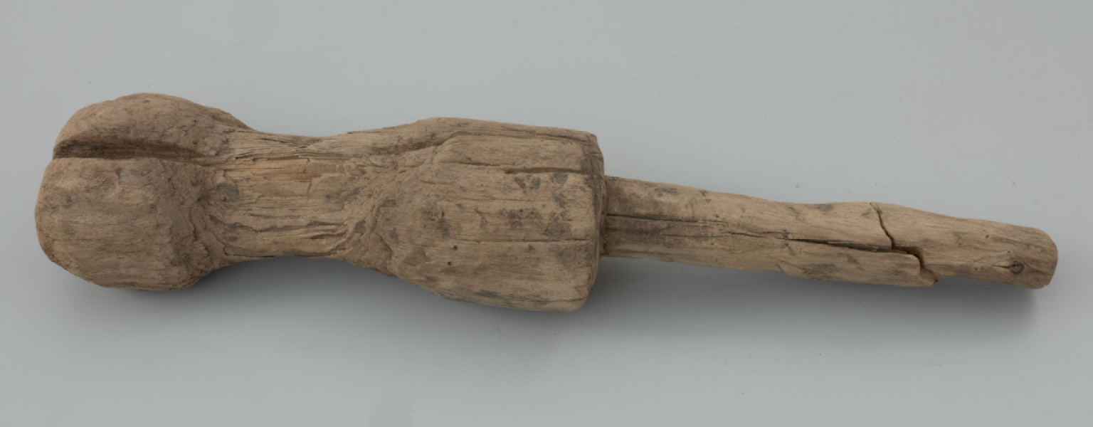 pałka ciesielska - Ujęcie z przodu. Przedmiot składa się z owalnego trzonka oraz elementu pracującego. Środkowa część zwana potocznie pobijakiem jest mocno wyeksploatowana z obustronnym łukowatym wgłębieniem. Na powierzchni zabytku możemy zaobserwować kilka interesujących śladów powstałych podczas jego produkcji. Są to między innymi podłużne ślady precyzyjnego strugania wykonane za pomocą ostrego noża, jak również lekkie wgłębienia świadczące o użyciu żelaznego ciosła.