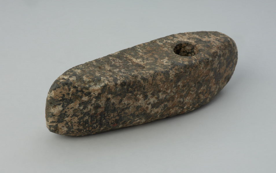 Topór kamienny - Ujęcie z tyłu skosem w lewą stronę; Smukły, mały topór z otworem umieszczonym w części obuchowej. Korpus lekko asymetryczny, z ostrzem minimalnie odgiętym w dół. Zarys zbliżony do pięciobocznego, z wąskim, tępym obuchem. Otwór mały, kolisty, stożkowaty, przechylony na jedną stroną (u dołu bliżej jednej krawędzi bocznej). Powierzchnia szlifowana, najsłabiej na stronie spodniej.