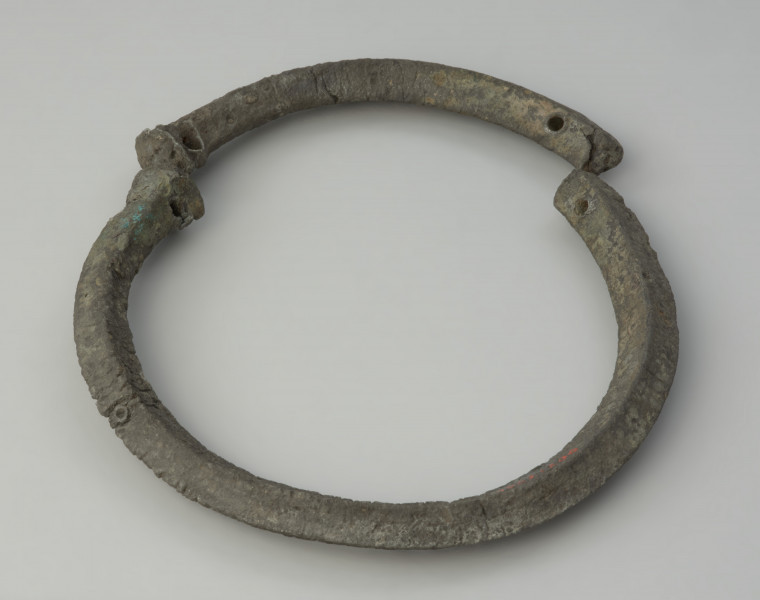 obręcz z czworograniastej sztaby - Ujęcie przodu obręczy z góry. Srebrzystoszara obręcz, rozłamana na dwie części, wykonana została z czworograniastej cynowej sztaby. Przypomina kształtem naszyjnik obręczowy z ściętymi prosto i nieco pogrubionymi końcami oraz niewielkimi, nieregularnie rozmieszczonymi otworami. Zabytek jest zdeformowany, a jego powierzchnia popękana, co może świadczyć o wtórnym przepaleniu w ogniu.