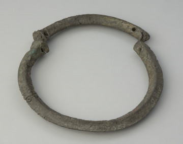 obręcz z czworograniastej sztaby - Ujęcie przodu obręczy z góry. Srebrzystoszara obręcz, rozłamana na dwie części, wykonana została z czworograniastej cynowej sztaby. Przypomina kształtem naszyjnik obręczowy z ściętymi prosto i nieco pogrubionymi końcami oraz niewielkimi, nieregularnie rozmieszczonymi otworami. Zabytek jest zdeformowany, a jego powierzchnia popękana, co może świadczyć o wtórnym przepaleniu w ogniu.