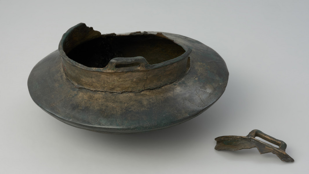 misa do zawieszania - Ujęcie z boku. Misa o dwustożkowatym brzuścu, przechodzącym w niską cylindryczną szyjkę z dwoma prostokątnymi uchami przy krawędzi. Dno naczynia uformowane jest kuliście, jego podstawa zaznaczona jest pojedynczym motywem koncentrycznych okręgów wykonynych w formie plastycznych żeberek. Jest to zarazem punkt centralny całej dekoracji dolnej części naczynia, poniżej załomu brzuśca. Punkt centralny wpisany jest w cztery okręgi. Pierwszy, najmniejszy tworzy wybijany motyw promienisty. Drugi, to podwójne pasmo wybijanych punktów a między nimi pasmo ryte złożone z dwóch linii, połączonych drobnymi ukośnymi nacięciami. Trzeci okrąg składa się ośmiu kolistych koncentrycznych żeberek, analogicznych do żeberek w punkcie centralnym naczynia, ale mniejszych. Ostatni okrąg tworzy potrójne żebrowanie przy krawędzi załomu brzuśca naczynia, połączone z łukowatymi festonami rytymi i wybijanymi. Górną część brzuśca zdobi natomiast linia falista (zygzakowa), także punktowana.