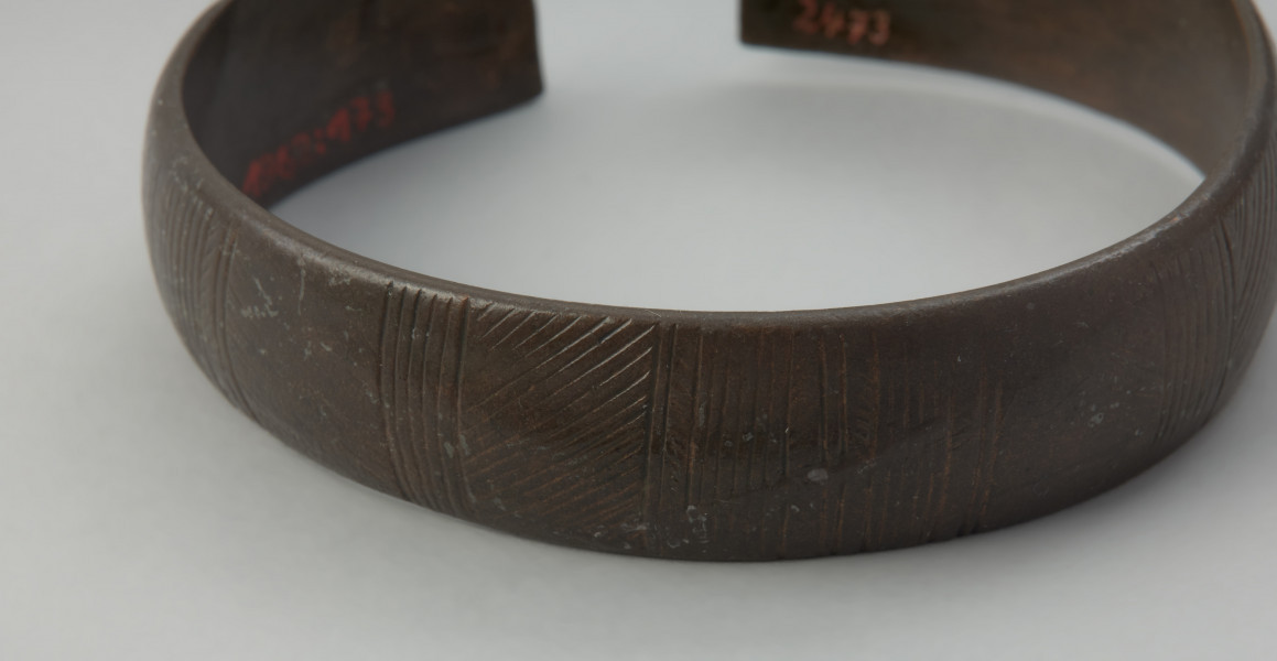 bransoleta mankietowata - Ujęcie z przodu. Bransoleta, tworząca szeroką otwartą obręcz, wykonana jest z blachy brązowej. Przez całą długość środkowej części bransolety biegnie wąskie, wysklepione żeberko, które stanowi element dekoracyjny. Po obu jego stronach umieszczono ornament w postaci rytej jodełki, prostokąta i niewielkich łuków.