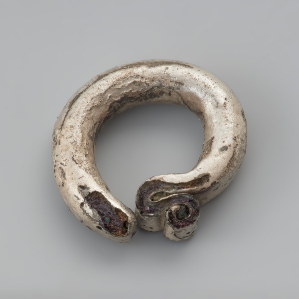 kabłączek skroniowy - Ujęcie dołu z góry. Lity kabłączek skroniowy wykonany z drutu o okrągłym przekroju poprzecznym z esowatym, spłaszczonym uszkiem podwójnie zagiętym. Powierzchnia pokryta została srebrną powłoką, obecnie jest spatynowana, nierówna, z kilkoma ubytkami, szczególnie przy końcach ozdoby.