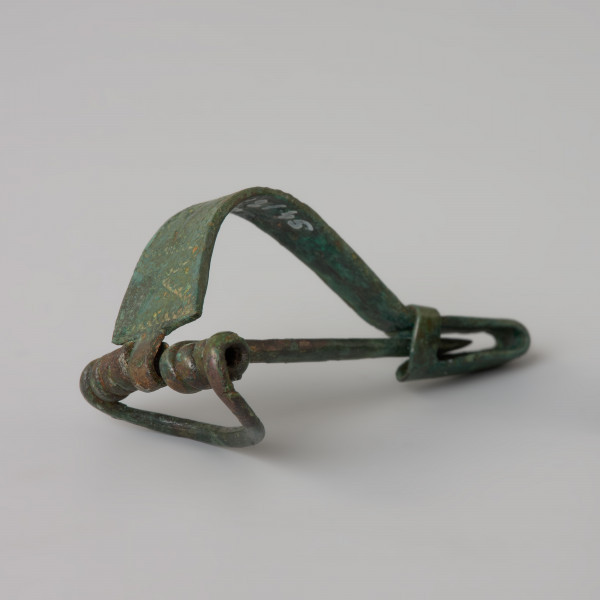 Fibula z brązu - Ujęcie z przodu z lewej strony. Fibula z brązu o podwiniętym na końcu kabłąku uformowanym w zaczep na szpilę (nóżkę). Zdobiona zygzakami na kabłąku.