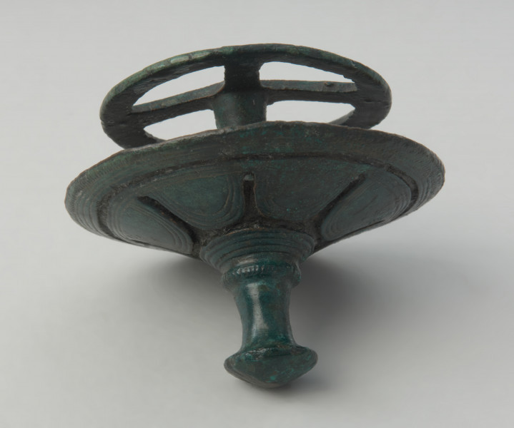 Brązowy tutullus złożony z dwóch tarczek oraz kolca. - Ujęcie tyłu tutullusa w pionie. Brązowy tutullus złożony z dwóch tarczek oraz kolca. Górna tarczka posiada  zdobienie w postaci żłobków, ażuru, linii rytych, nacięć oraz motywu solarnego. Dolna tarczka ma formę koła z czterema szprychami.