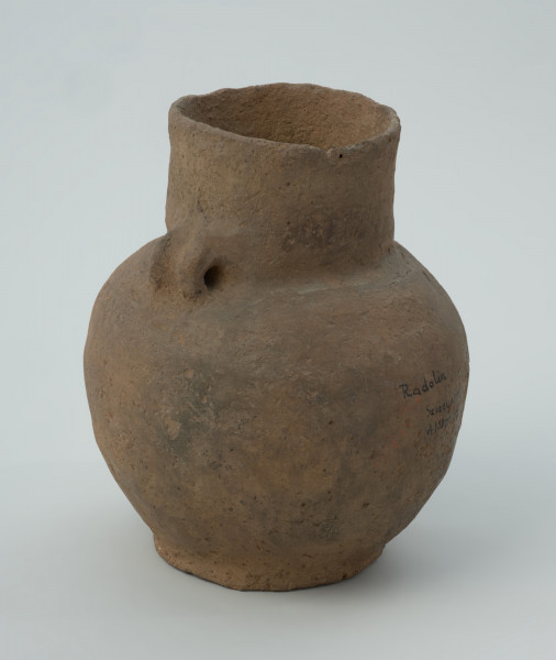 ceramika, naczynie - Ujęcie z boku; Niezdobiona amfora z niemal cylindryczną szyjką, z parą taśmowatych, wątorowanych uszek na załomie szyjki i brzuśca. Z kulistym brzuścem i niską stopką. Uszkodzona przy przejściu szyjki w brzusiec (pęknięcie i brak ucha).