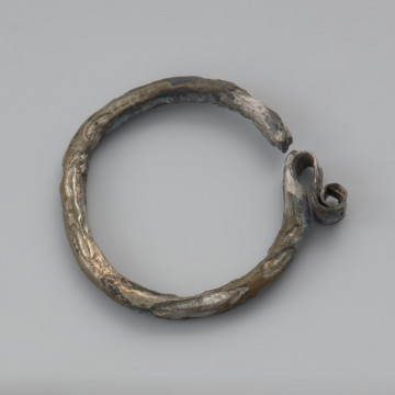 kabłączek skroniowy - Ujęcie dołu z góry. Brązowy kabłączek skroniowy z litego metalu z esowatym spłaszczonym uszkiem podwójnie zwiniętym w spiralę. Powierzchnia spatynowana, ciemnobrązowa.